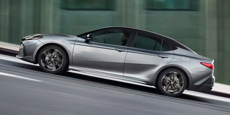 Silver sedan with black roof, side view, driving up a gray road at an angle with a green building blurring in the background.