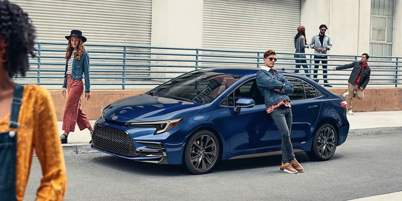 A dark blue sedan parked on a city street, with a man leaning on it. Several people are in the background.
