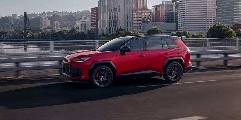 A red Toyota RAV4 driving on a highway, surrounded by green trees and clear blue skies.