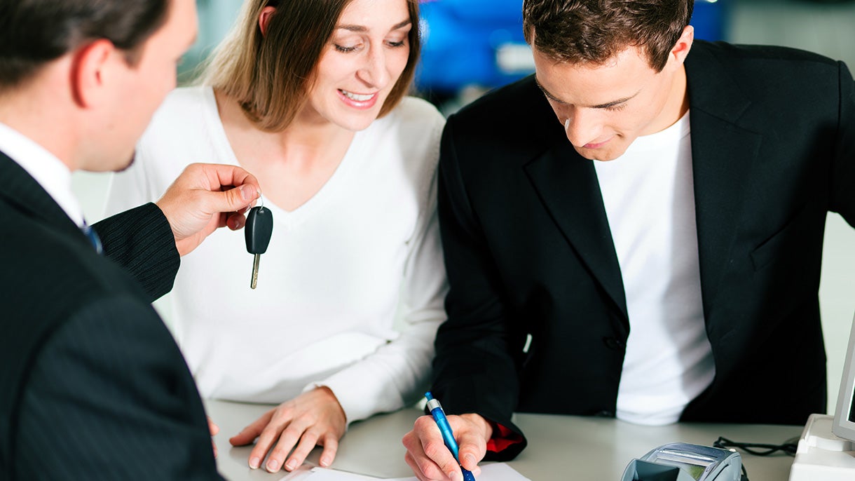 A couple and a car salesperson. The salesperson is handing over car keys to the smiling woman, whose partner is signing paper.