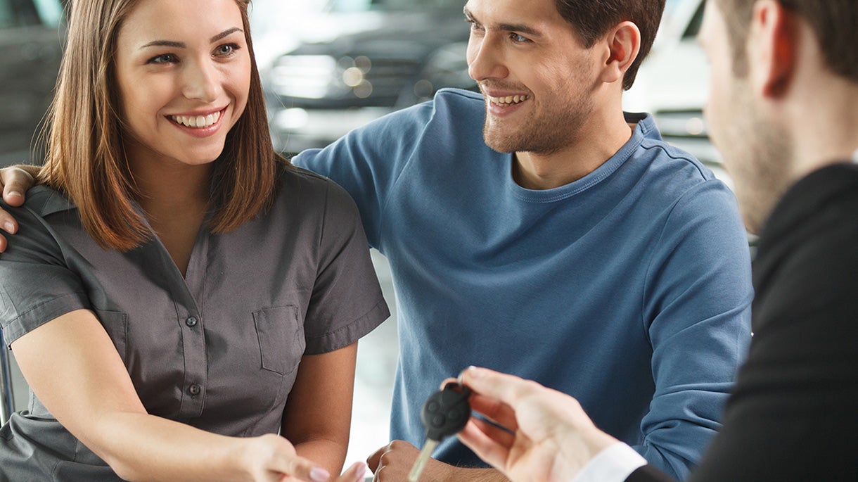A smiling couple receives a car key from a salesperson in a showroom.