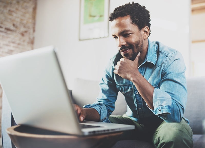 Smiling man with dark curly hair and beard, wearing a denim shirt, looking at a laptop while sitting on a couch.